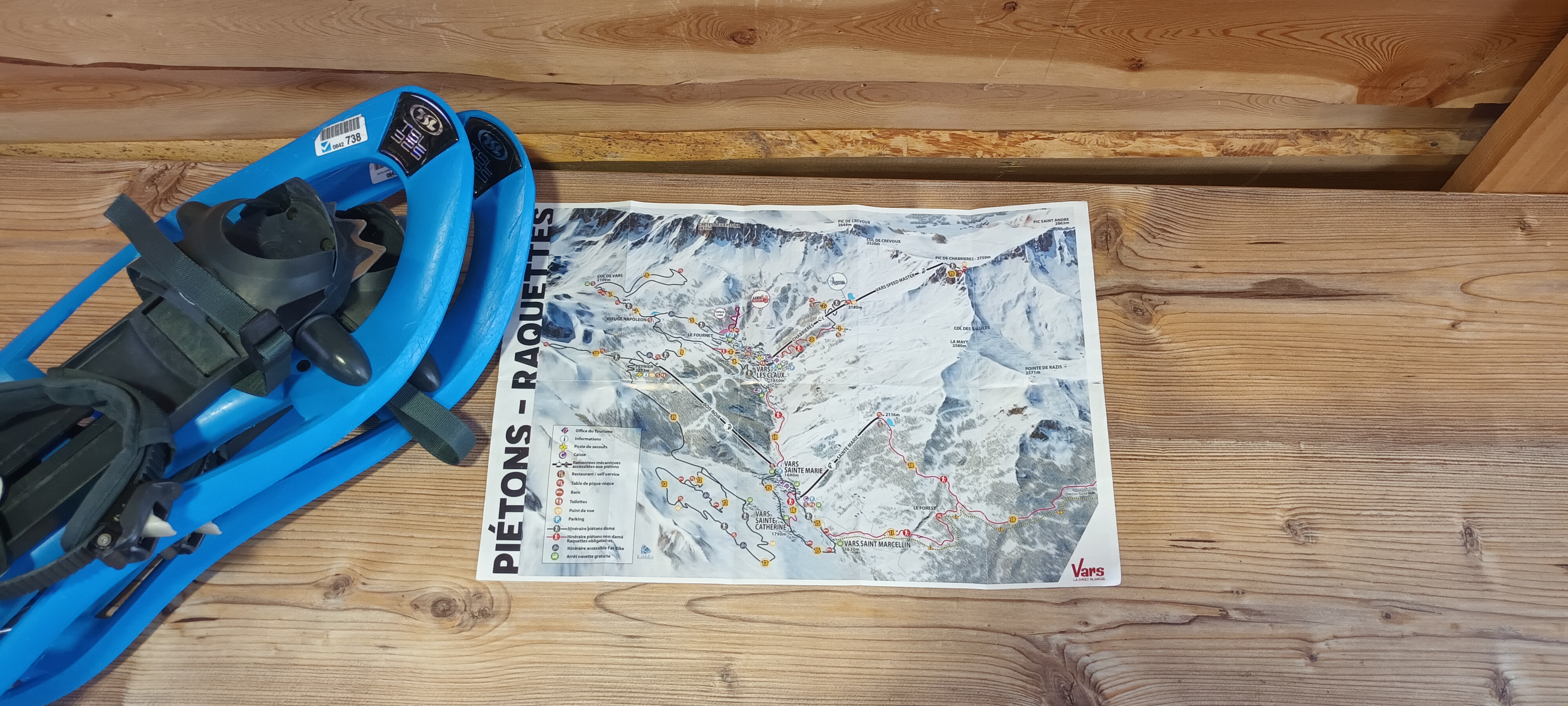 Pair of blue snowshoes and map of the hiking routes of the Vars resort on a wooden stand.
