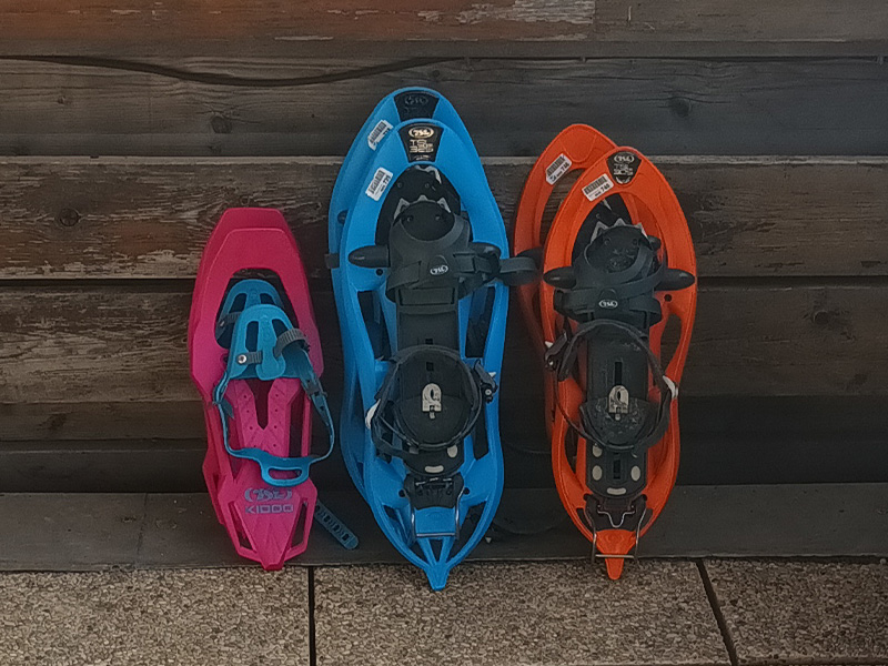 Three pairs of colorful snowshoes sitting on a terrace facing the snowy chalets of a ski resort.